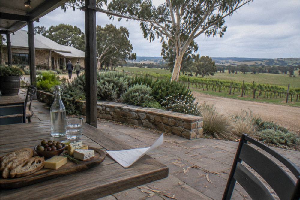 A shaded outdoor table at a Barossa winery with a platter of bread, cheese and olives and a bottle of water.