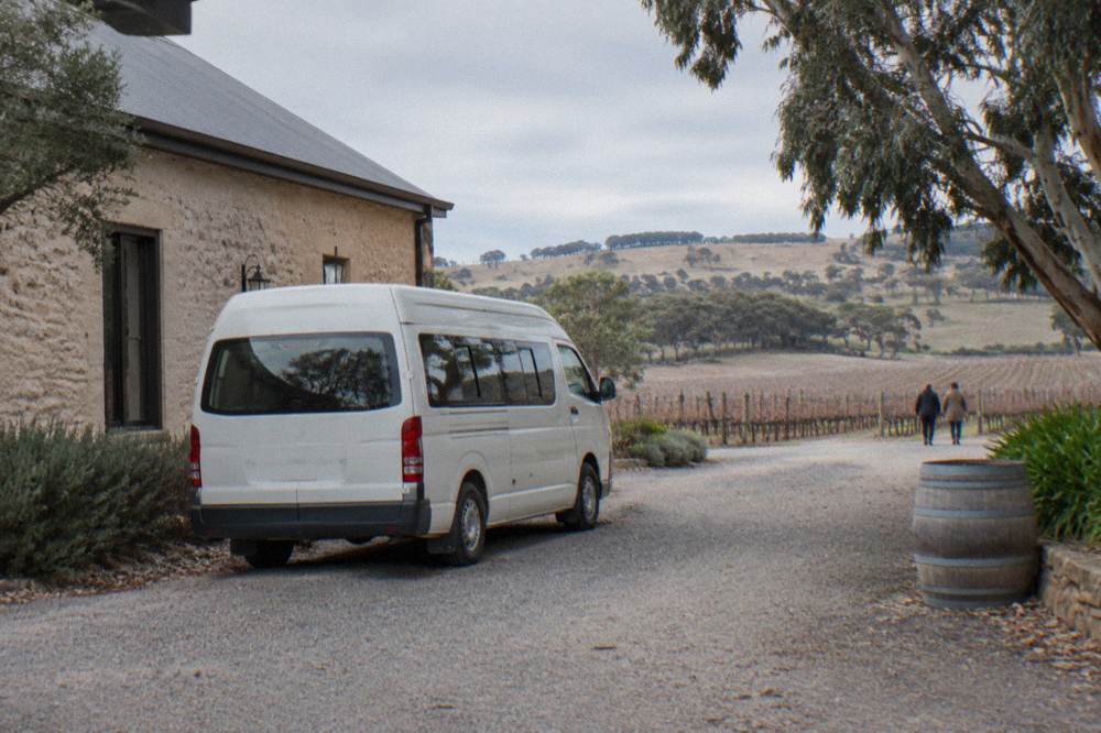 A small tour van parked on a gravel drive beside a stone winery building with no people