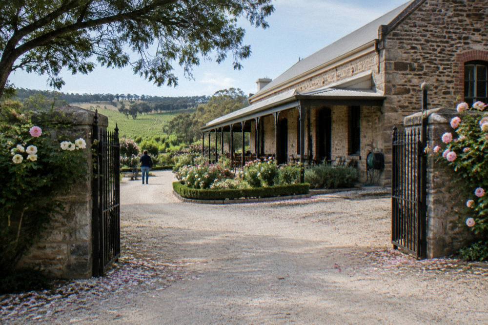 Stone winery building with a rose garden and iron gates at the end of a driveway