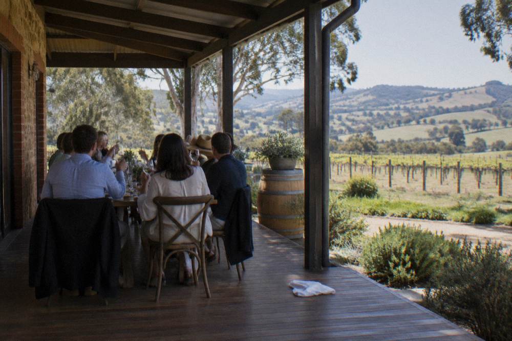 A group of people seated at a long table under a verandah, sharing platters of food and glasses of wine with a vineyard backdrop