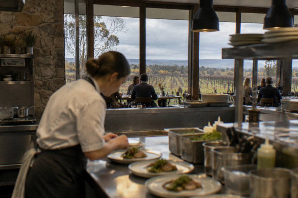 A chef plating a dish in an open winery kitchen with diners at tables behind, large windows showing vineyards