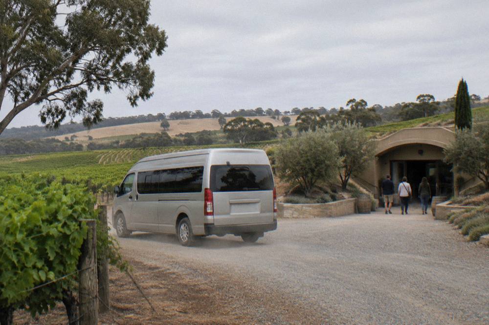 A small tour van parked on a gravel drive beside rows of vines and a cellar door entrance